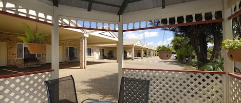 A covered outdoor seating area with a table and chairs, a hanging planter, and a view of a building with a covered walkway.