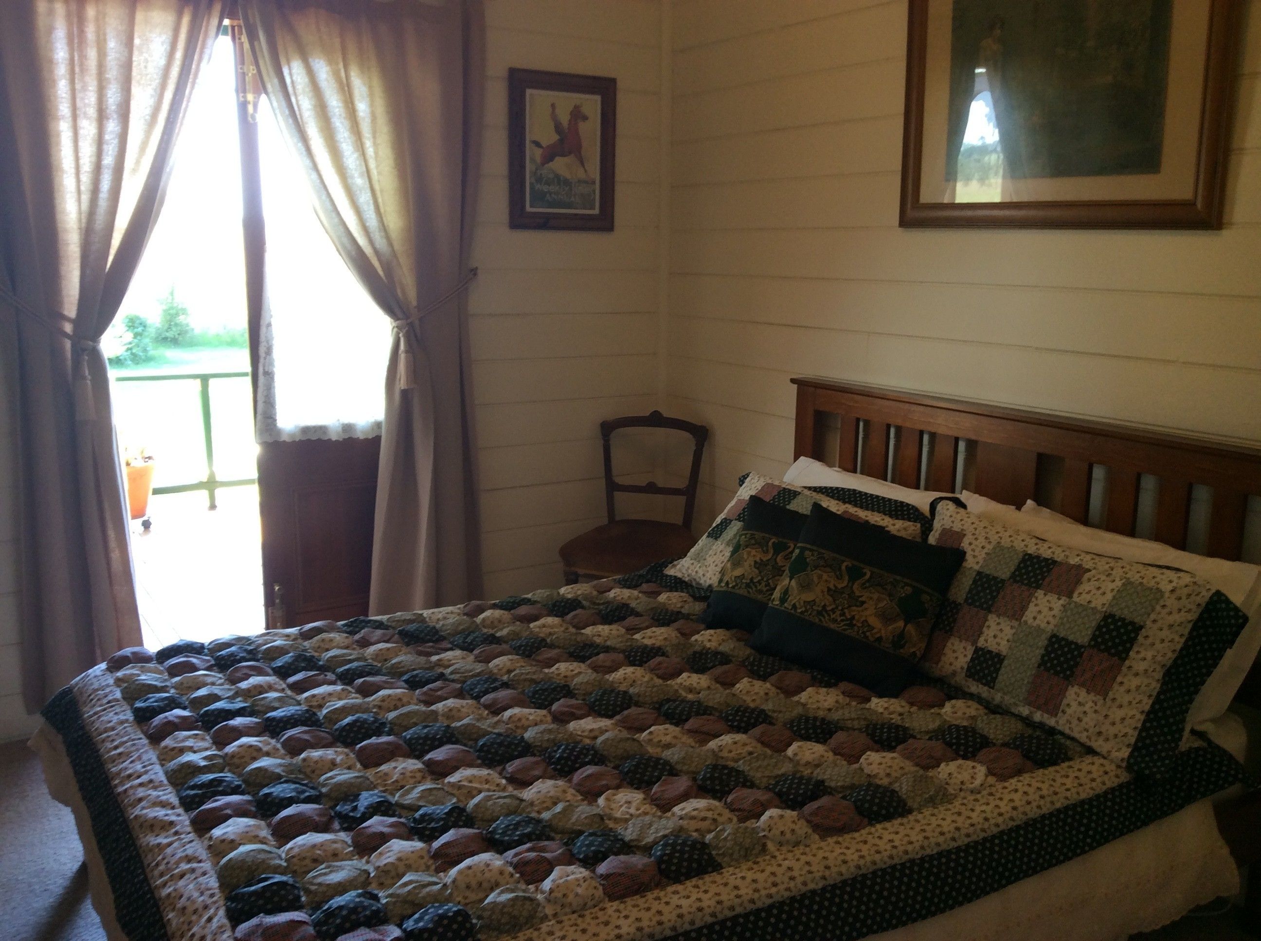 A bedroom with a wooden bed, a quilt, a chair, and a window with curtains.
