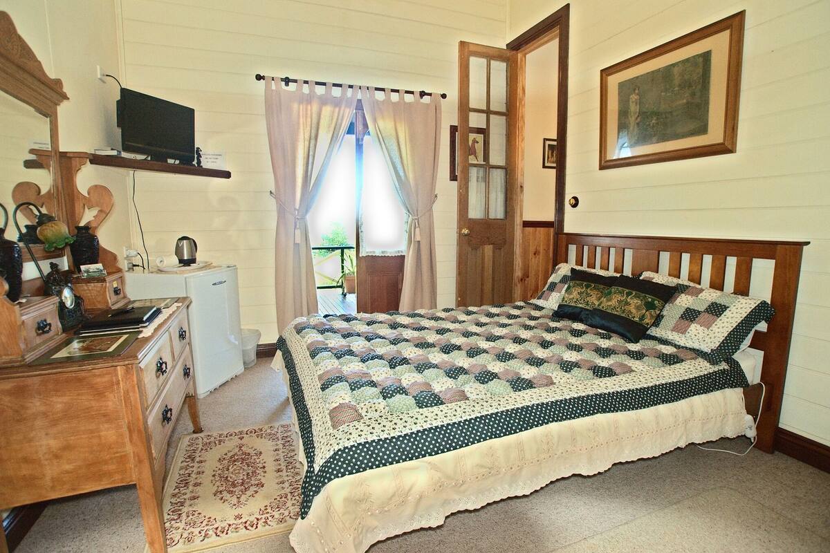 A bedroom with a wooden bed, a desk with drawers, a small cabinet, a television on a shelf, and a window with curtains.