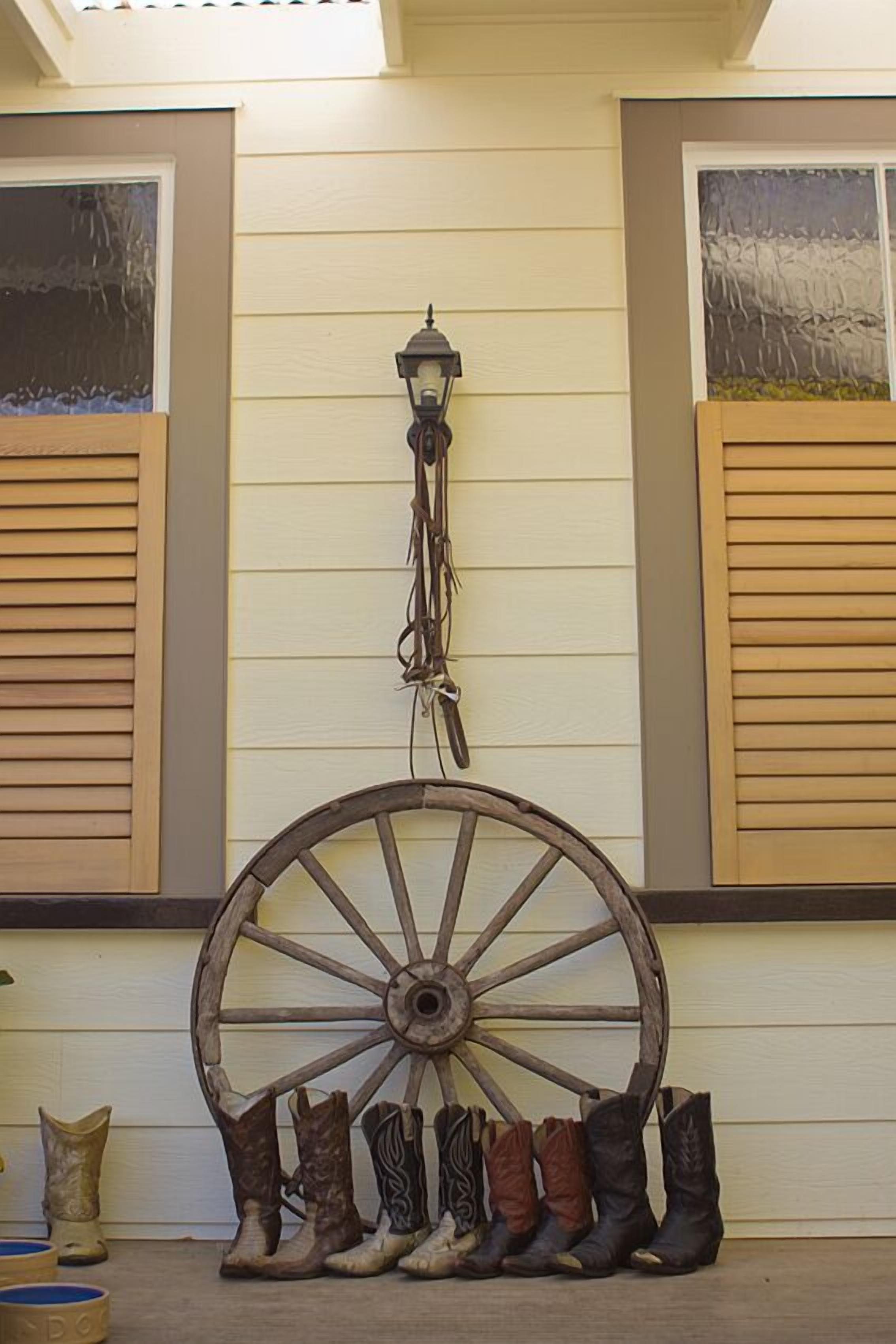Un décor extérieur rustique agrémenté d’une grande roue de wagon en bois, d’une lanterne et d’une rangée de bottes de cowboy, le tout adossé à un mur de couleur claire orné de volets.