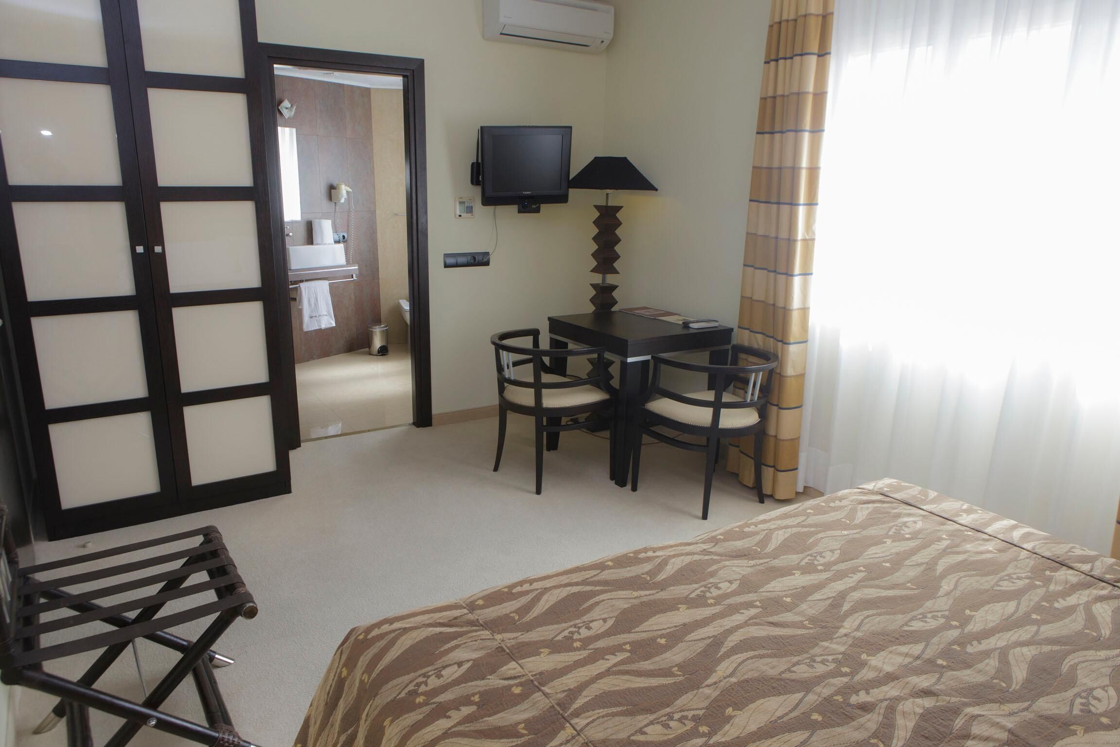 Une chambre d’hôtel comprenant un lit, un bureau avec une chaise, une télévision et une fenêtre avec des rideaux.