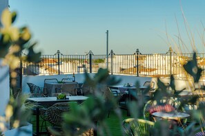 An outdoor patio with wicker furniture, a table with a bottle, and a view of buildings in the background. - San Miguel Hotel (Mahón)