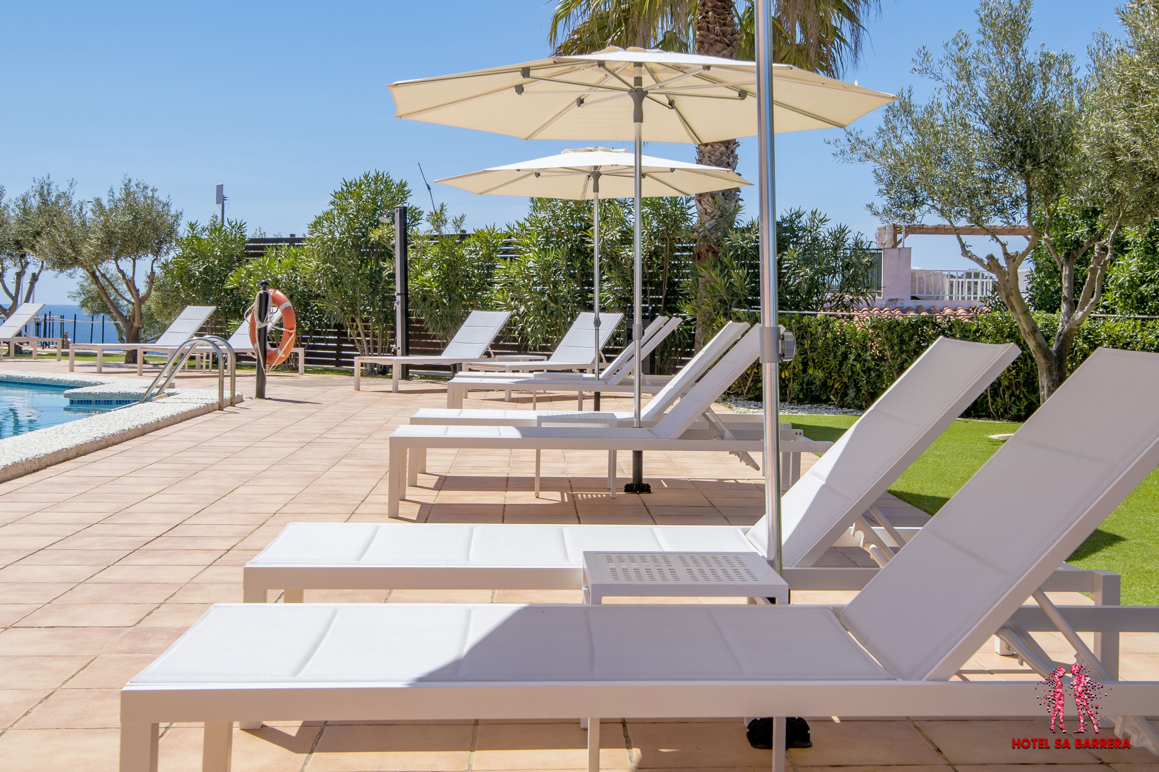 A poolside area with white lounge chairs and umbrellas, a lifebuoy, and a view of the sea.