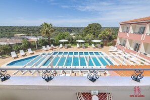 A rooftop pool with multiple lounge chairs and umbrellas, surrounded by lush greenery and a view of distant hills.