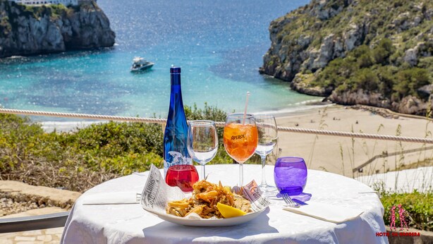 Una mesa con un plato de mariscos, dos copas de vino y una botella, con vistas a un paisaje costero con acantilados rocosos y una playa.