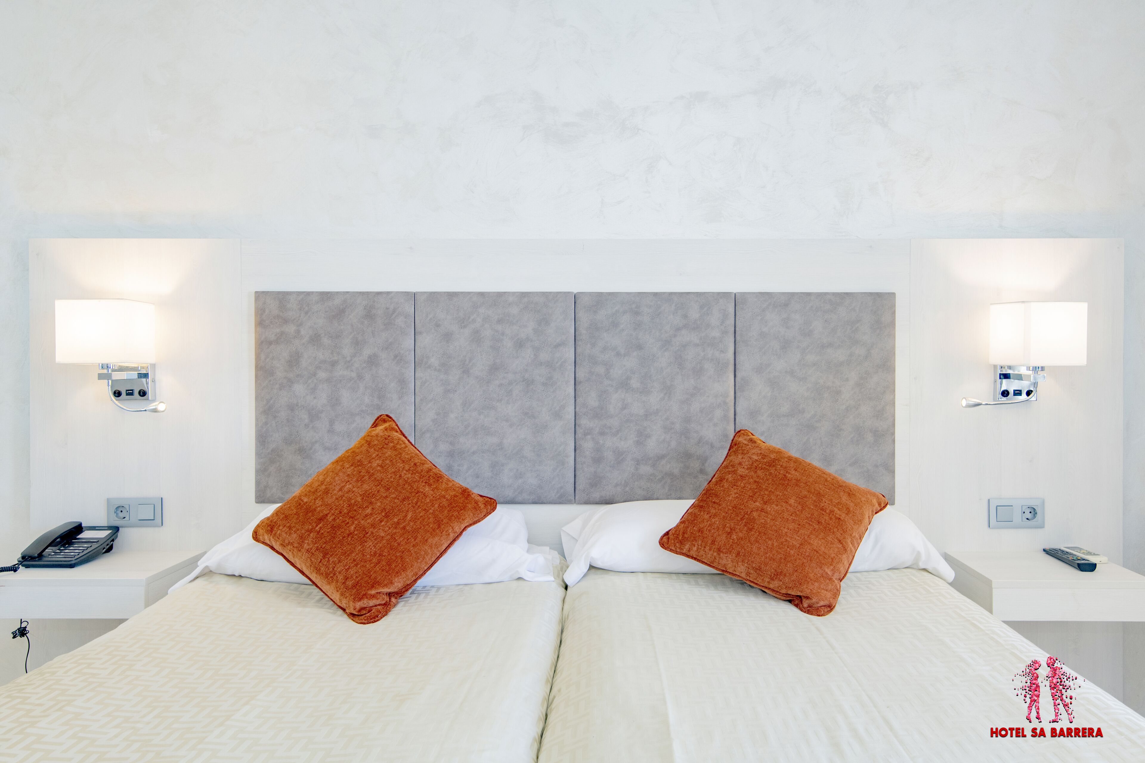 Ein Hotelzimmer mit zwei Betten, einem grauen Kopfteil, Nachttischlampen, einem Telefon und einer Fernbedienung.