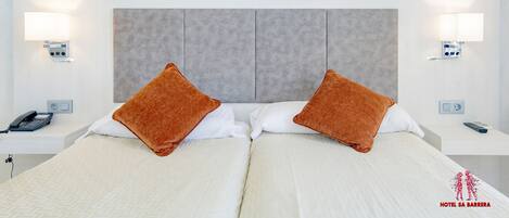 Ein Hotelzimmer mit zwei Betten, einem grauen Kopfteil, Nachttischlampen, einem Telefon und einer Fernbedienung.