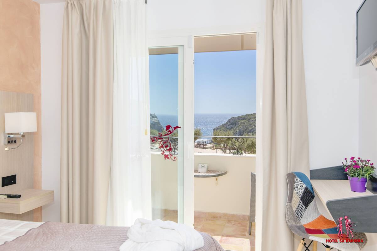 Une chambre d’hôtel avec un lit, un bureau, une chaise, un balcon donnant sur la mer et une plante en pot.