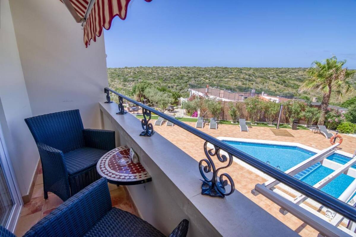 Un balcon agrémenté de meubles en osier, une piscine et une vue sur un espace paysager avec des arbres et un bâtiment en arrière-plan.