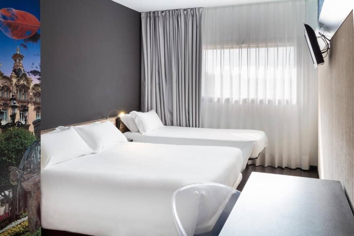 A hotel room with two beds, a desk, and a large window with curtains.