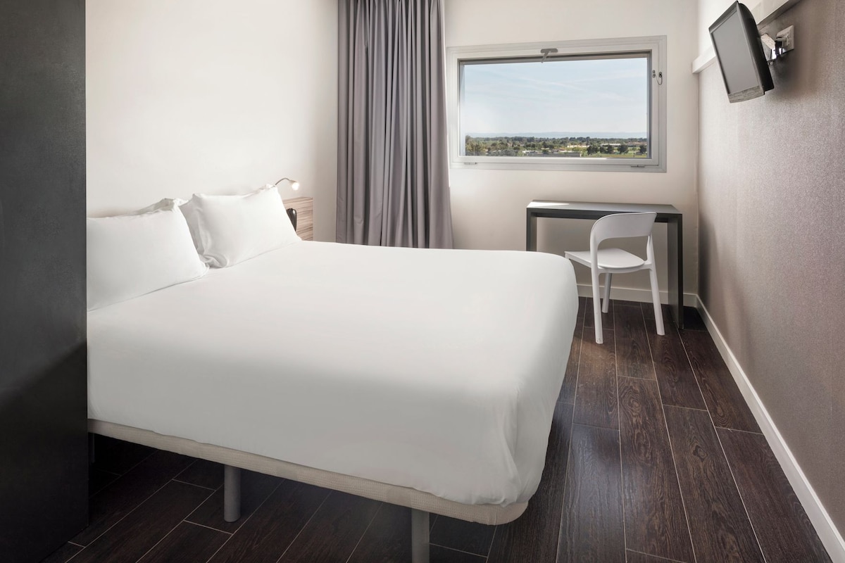 A hotel room with a bed, a desk, a chair, a window with a view, and a flat-screen TV mounted on the wall.
