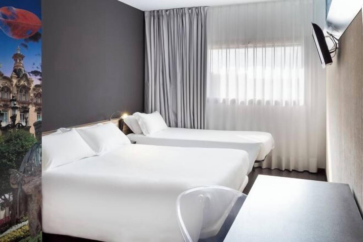 A hotel room with two beds, a large window with curtains, a flat-screen TV mounted on the wall, and a view of a cityscape.