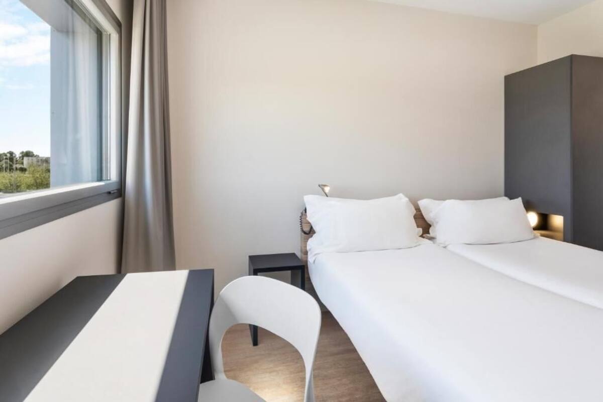 A hotel room with a bed, a desk, a chair, a nightstand, and a window.