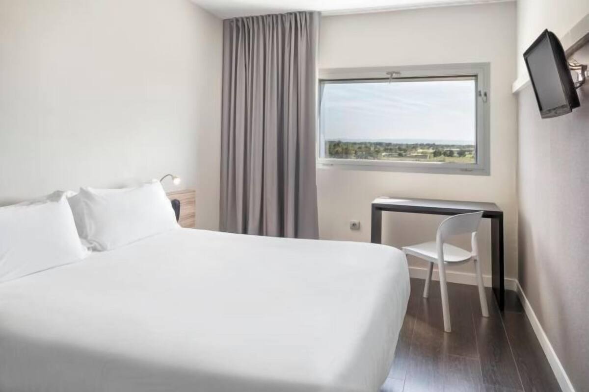 A hotel room with a bed, a desk, a chair, a TV, and a window with a view.