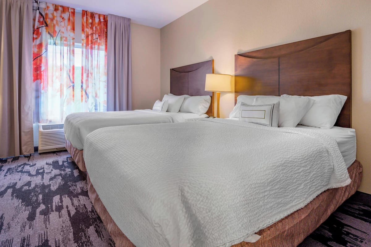 A hotel room with two beds, a large window with curtains, a wall-mounted lamp, and a patterned carpet.