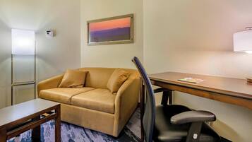 A hotel room with a beige sofa, a wooden desk, a black office chair, a framed painting on the wall, and a window with blinds.