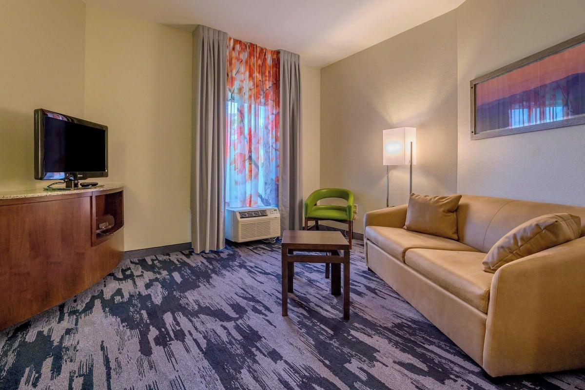 A hotel room with a beige sofa, a green chair, a television on a wooden stand, a lamp, and a window with curtains.