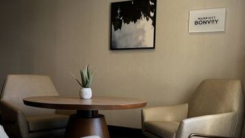 A hotel room with a round wooden table, two beige armchairs, a potted plant, a framed artwork, and a sign reading "MARRIOTT BONVOY".