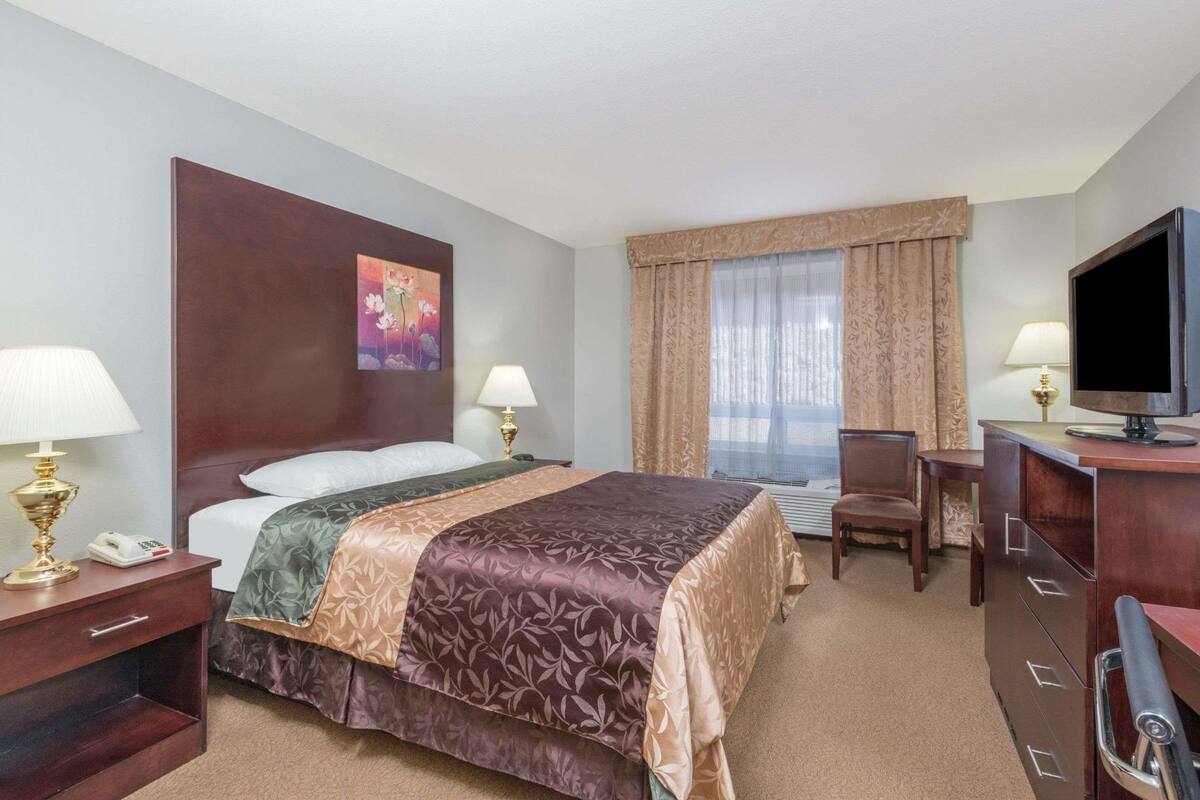 A hotel room with a bed, bedside lamps, a desk with a chair, a television, and a painting on the wall.