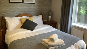 A neatly made bed with white and gray bedding, a wooden headboard, and a nightstand with a lamp and a bottle. - The Bolingbroke Hotel (Swindon)
