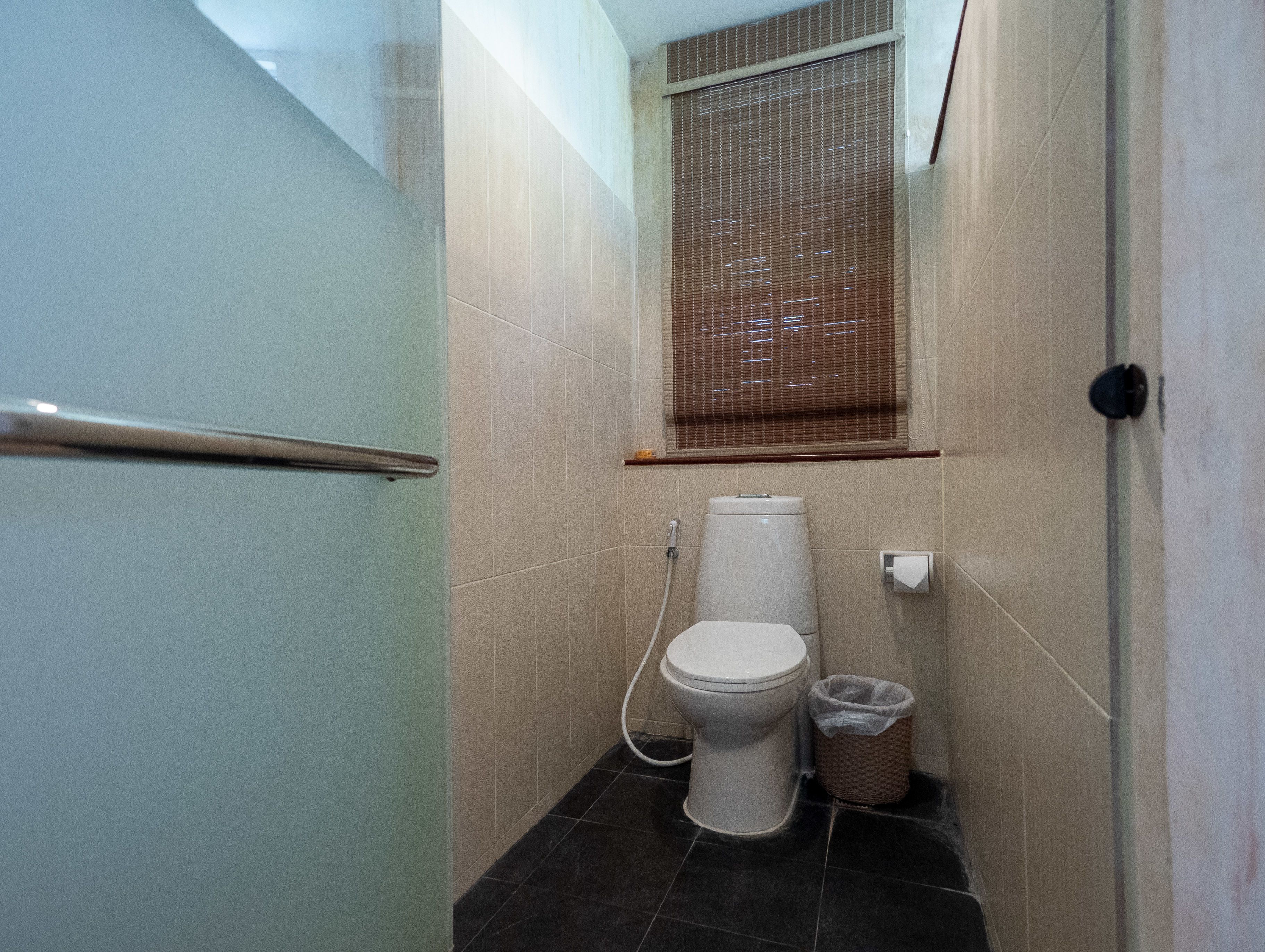 A small, narrow bathroom with a toilet, a trash bin, and a window with blinds.