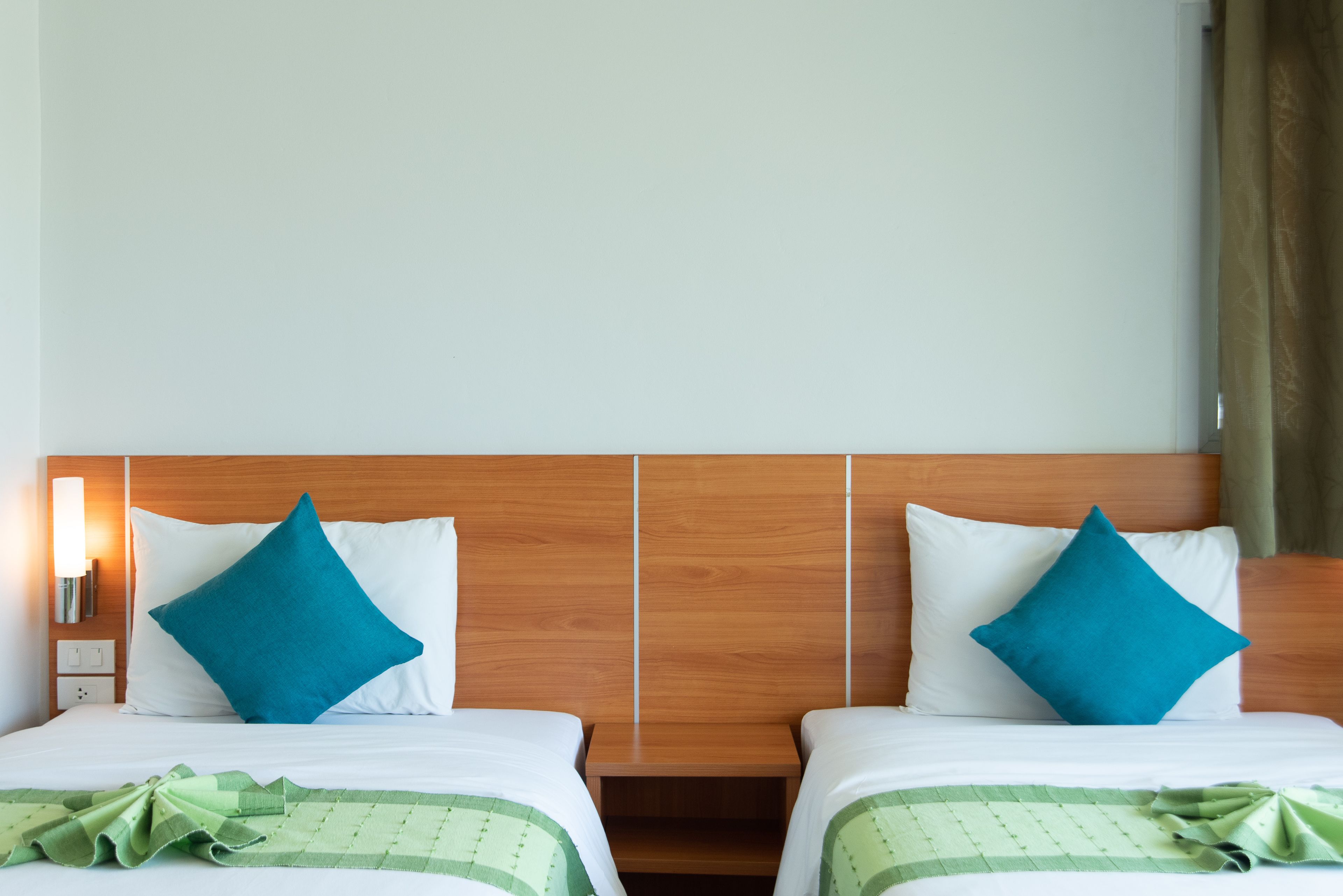Ein Hotelzimmer mit zwei Betten, jeweils mit einem Holz-Kopfteil, weißer Bettwäsche und türkisblauen Kissen. Auf einem Bett liegt eine grün karierte Decke.