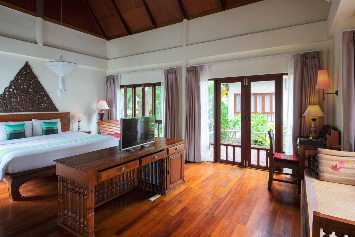 A spacious room with a large bed, wooden furniture, and a television. There is a balcony with a view of greenery.