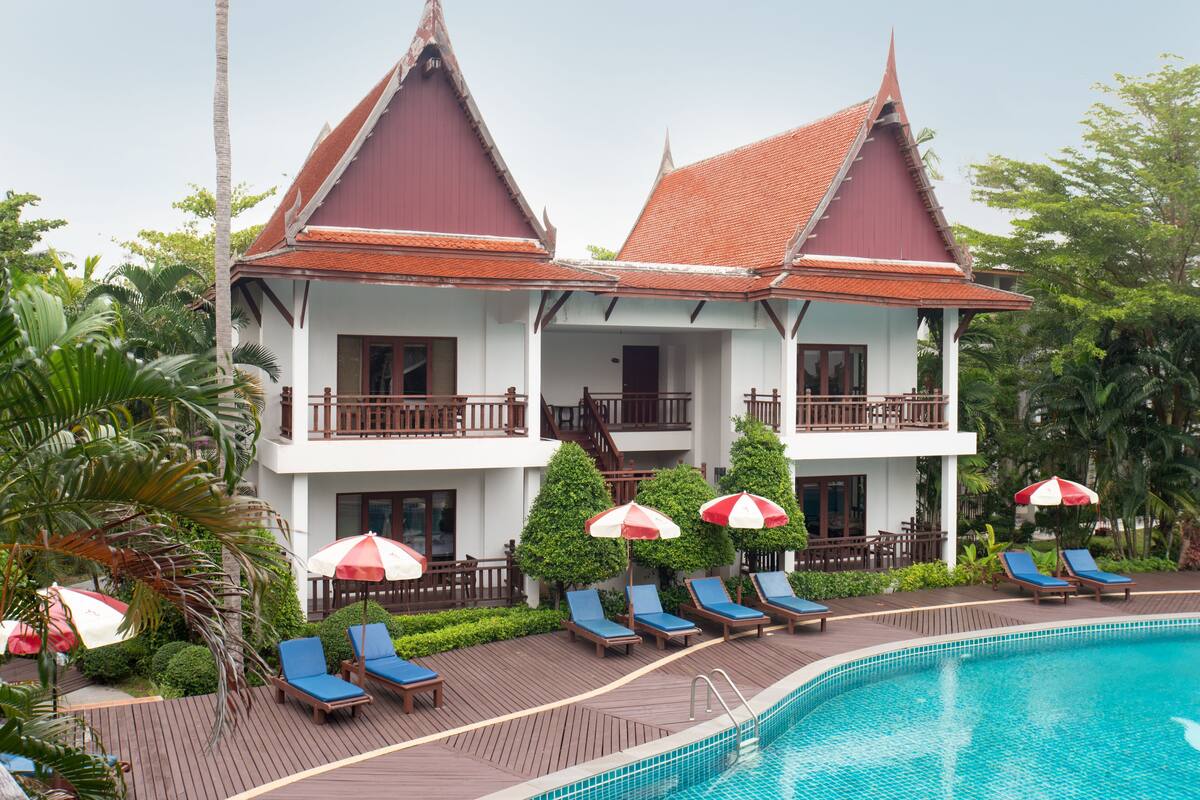 A two-story building with a red roof and white walls, a swimming pool, and lounge chairs with umbrellas.