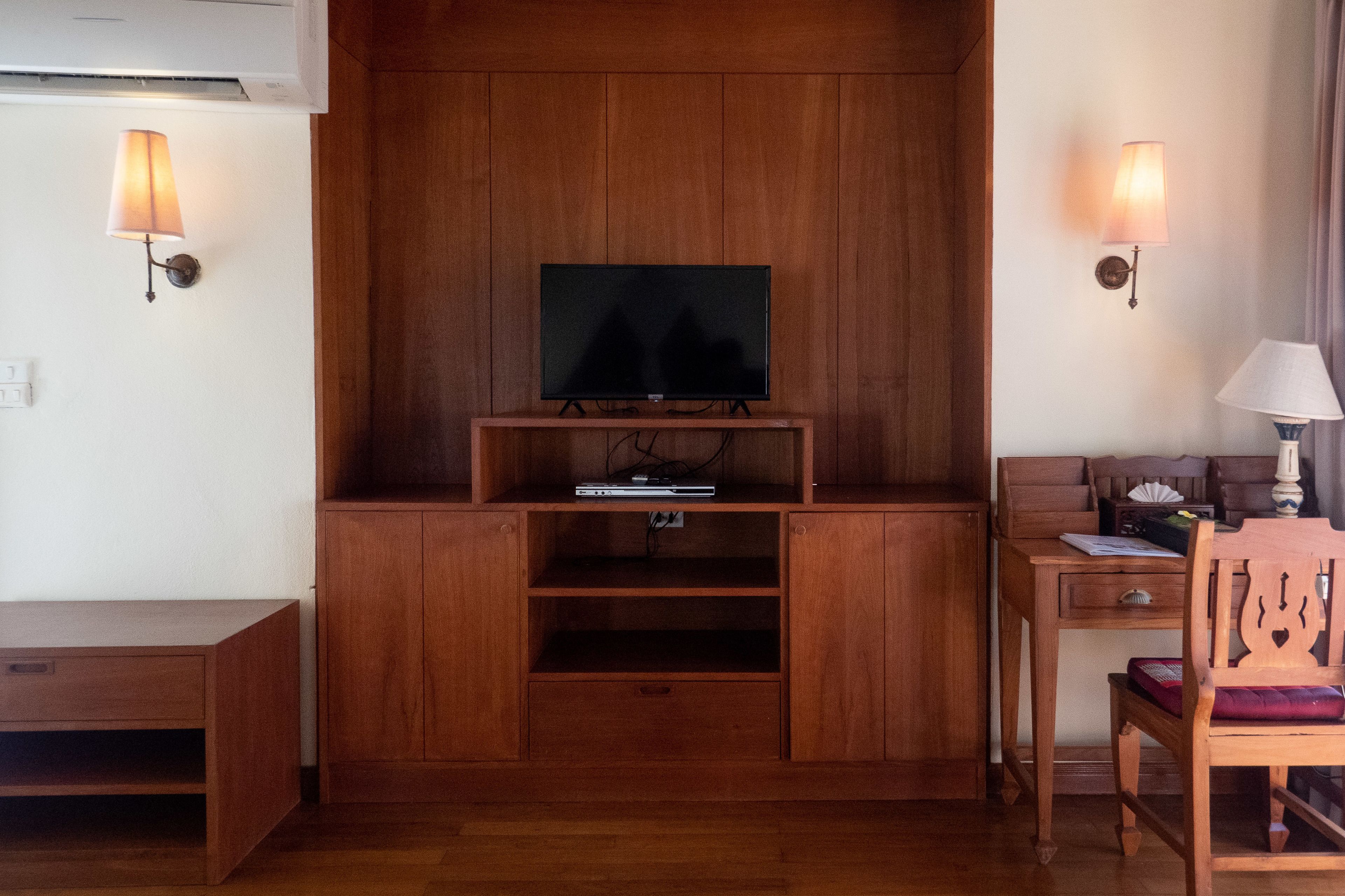 A wooden entertainment unit with a television, two wall-mounted lamps, and a desk with a chair.