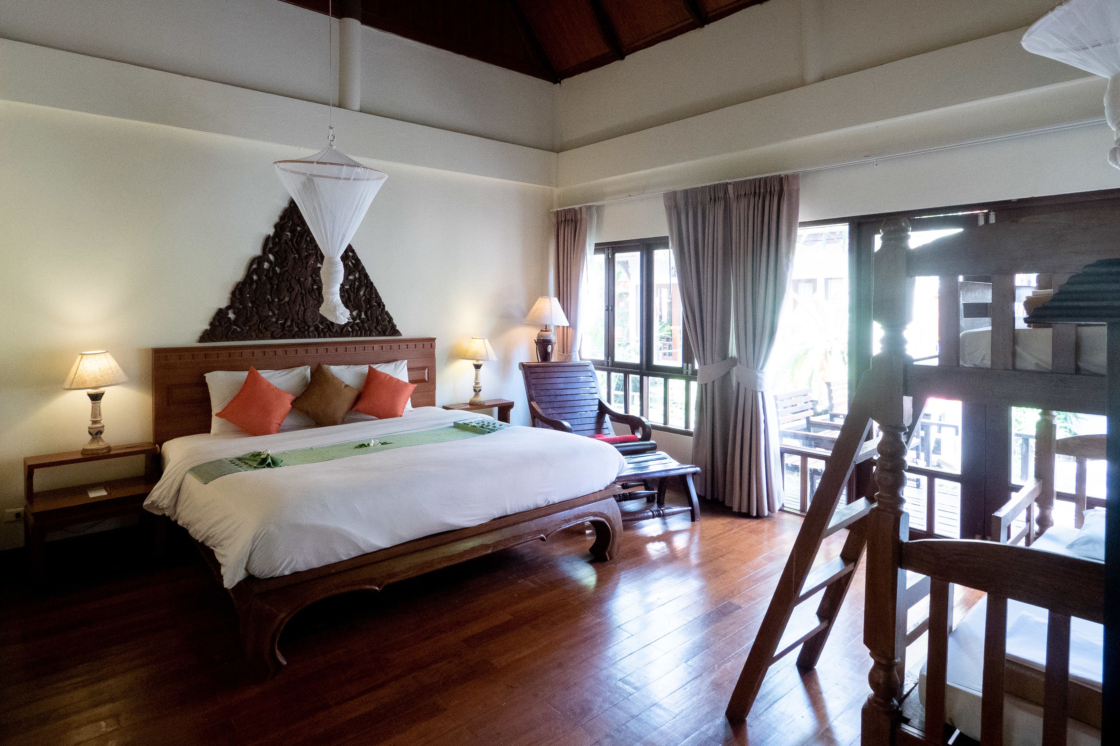 A bedroom with a wooden bed, two bedside tables with lamps, a chair, and a view through large windows.
