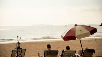 A beach scene with a row of beach chairs and an umbrella, a lamp post, and a distant view of the sea and a sailboat.