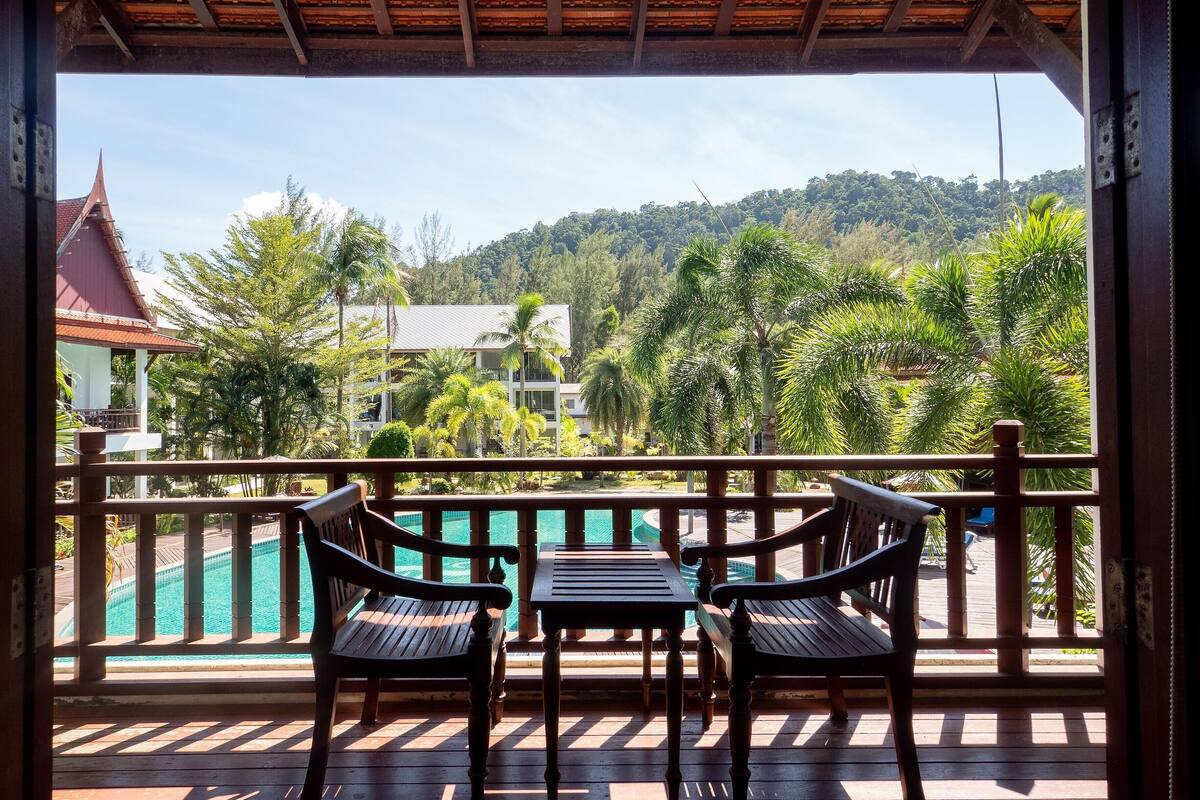 A wooden deck with two chairs and a table overlooking a swimming pool and a view of a building with a red roof.