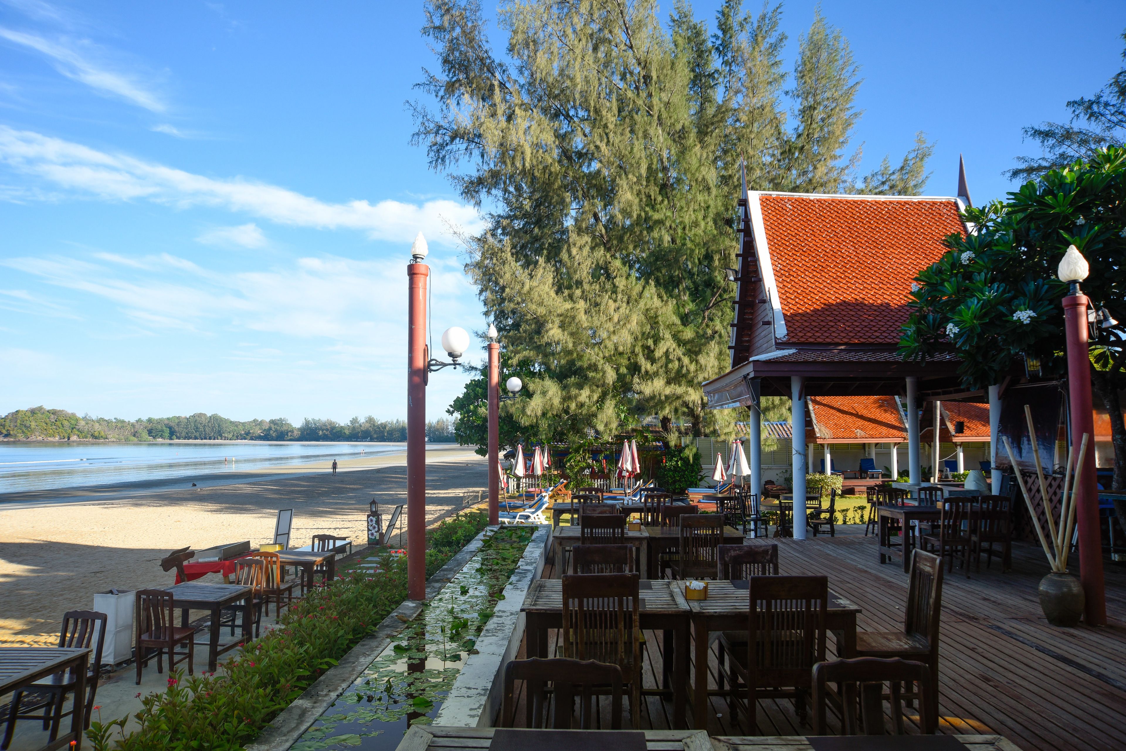 An outdoor dining area with wooden tables and chairs, a view of the beach, and a building with a red roof.