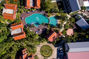 Vista aérea de un resort con piscina central, palmeras y edificios con tejados rojos.