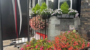 Un bâtiment avec une enseigne indiquant « Brantfell » et une affiche « COMPLET » sur la fenêtre. On y voit des jardinières remplies de différentes plantes et fleurs, ainsi qu’une clôture en métal ornée de fleurs roses au premier plan.