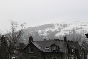 A stone building with multiple chimneys and a sloped roof, surrounded by trees and a foggy background. - Brantfell House (Ambleside)