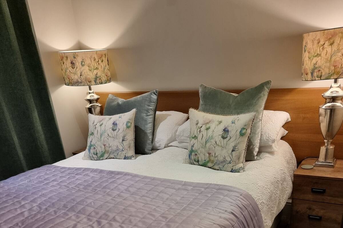 A neatly made bed with a purple quilt, floral pillows, and a wooden headboard. There are two bedside lamps with floral lampshades and a small wooden nightstand with drawers.