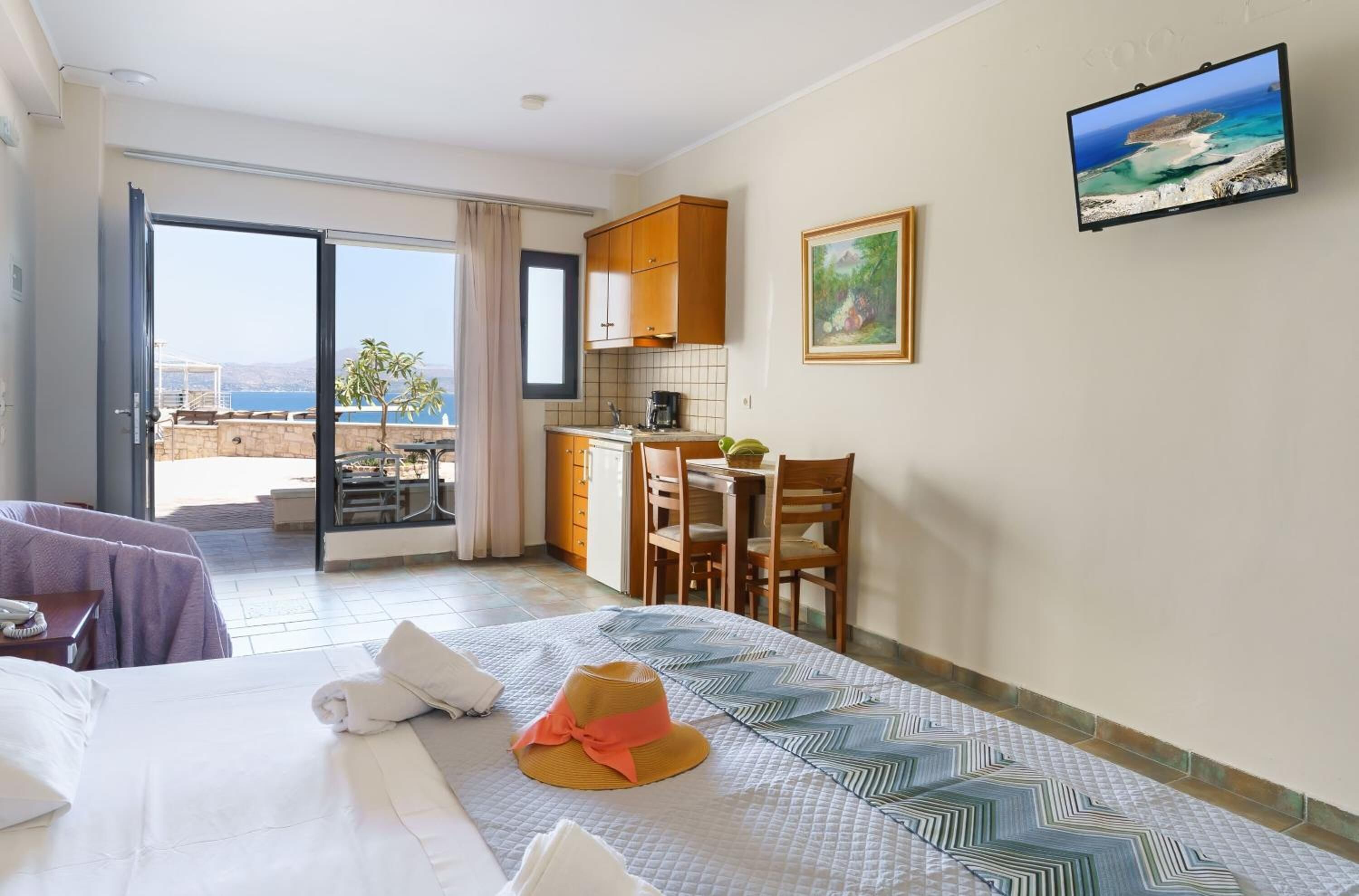 A hotel room with a bed, a dining table with chairs, a kitchen area, and a balcony with a view of the sea.