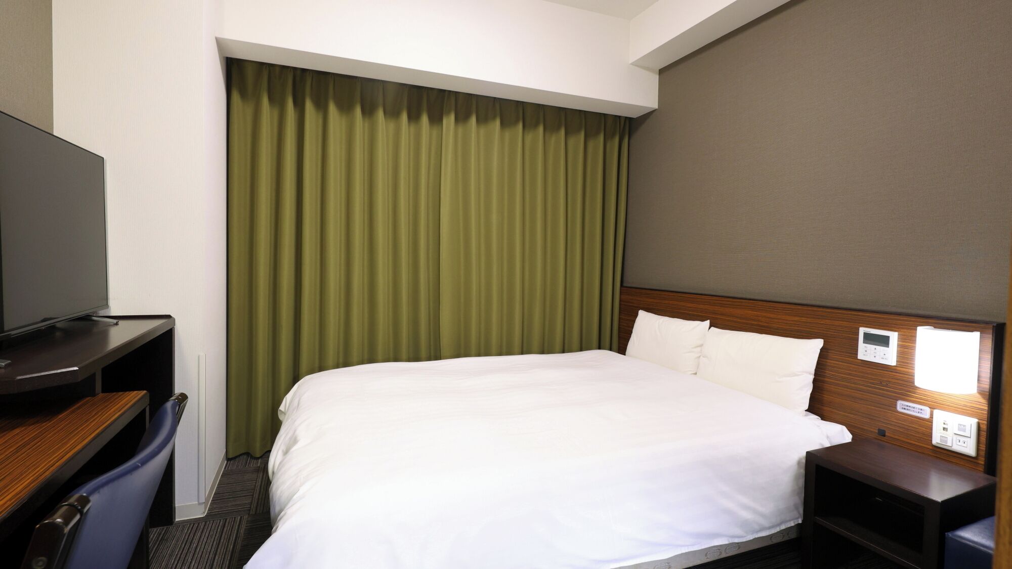 A hotel room with a bed, a desk, a television, and a control panel on the wall.