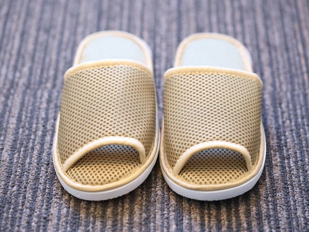 A pair of beige mesh slippers with white soles on a textured surface.