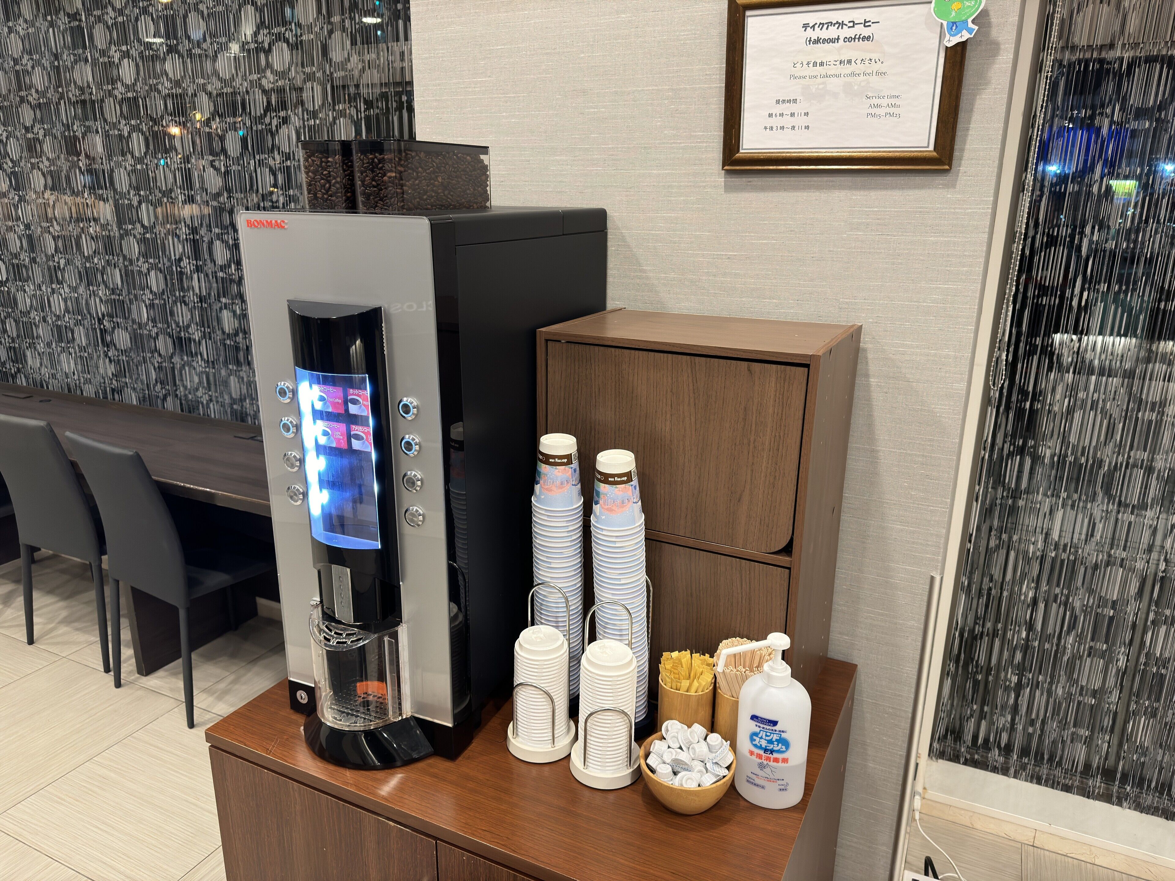 Une machine à café dotée d’un écran numérique, une pile de gobelets en papier, une bouteille de gel hydroalcoolique et un panneau encadré posé sur un comptoir.