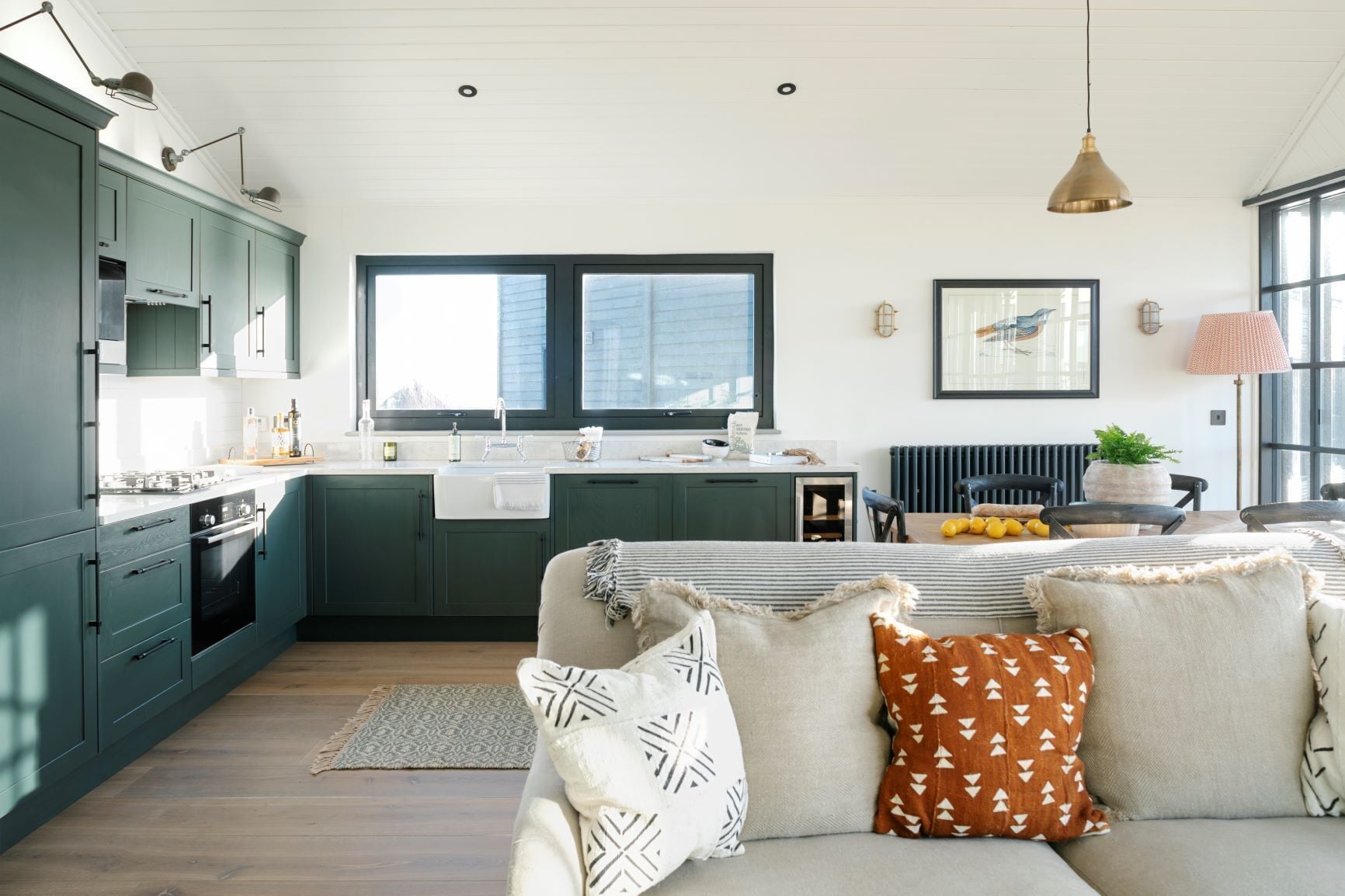 A modern kitchen with dark green cabinets, a large window, and a dining area with a sofa and cushions.