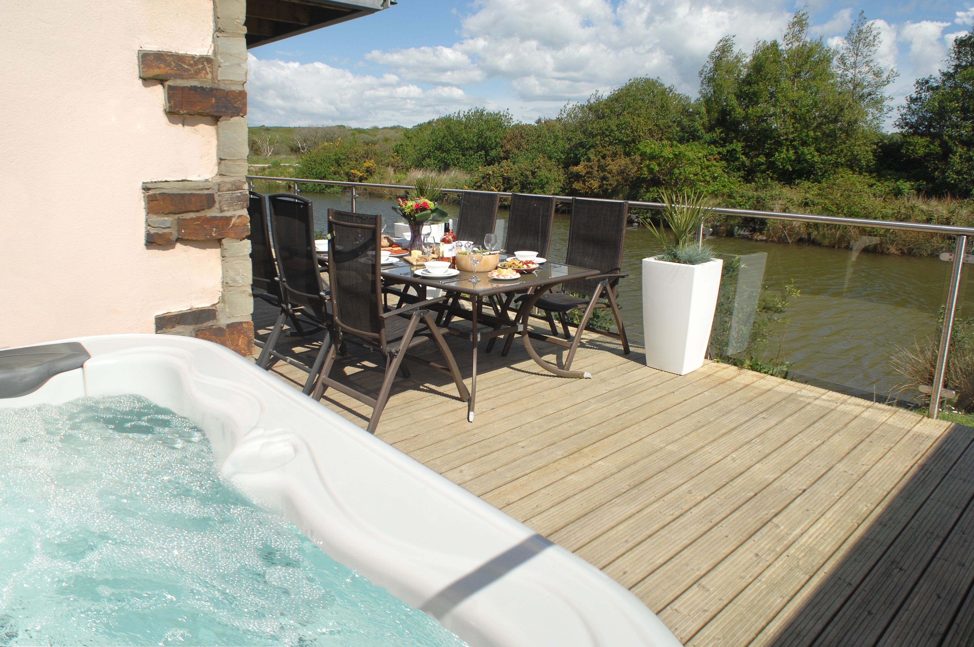 A wooden deck with a hot tub, dining table, and chairs overlooking a body of water.