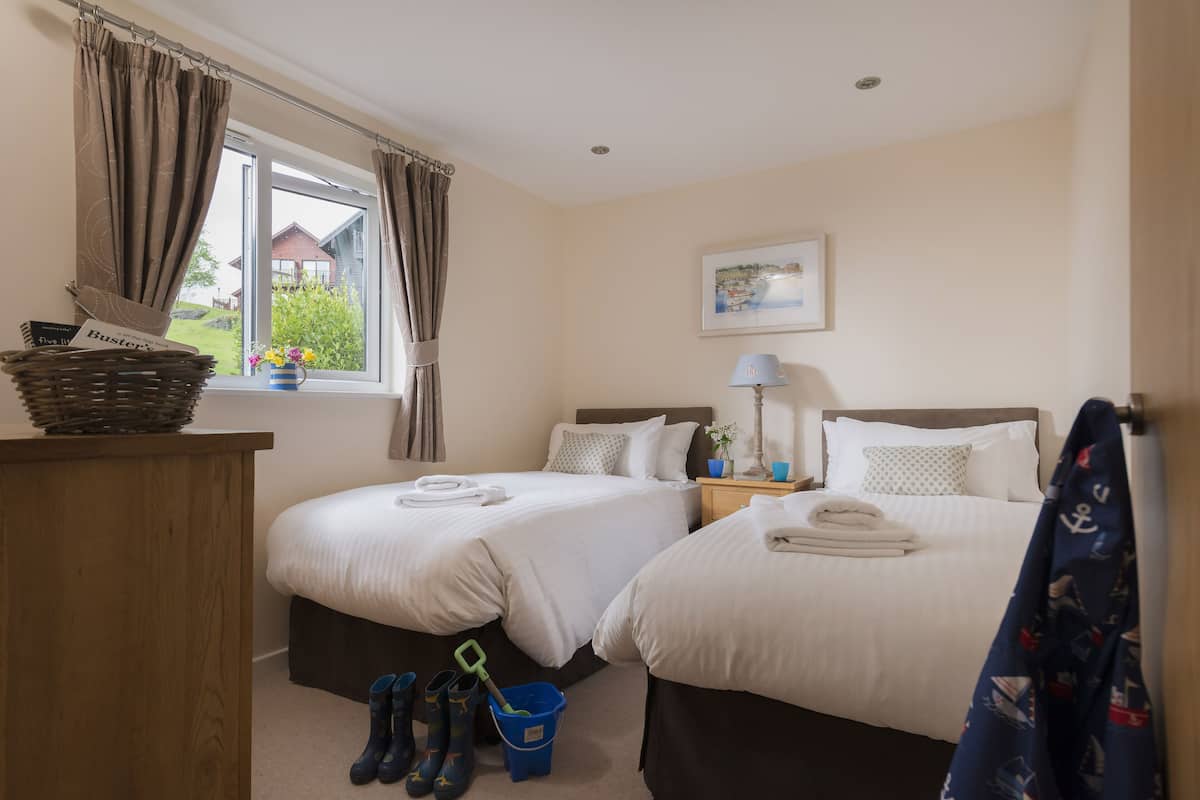A hotel room with two beds, a window with curtains, a painting on the wall, a lamp on the nightstand, and a basket on the dresser.