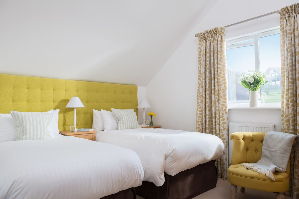 A hotel room with two beds, a yellow headboard, a yellow armchair, and a window with curtains.
