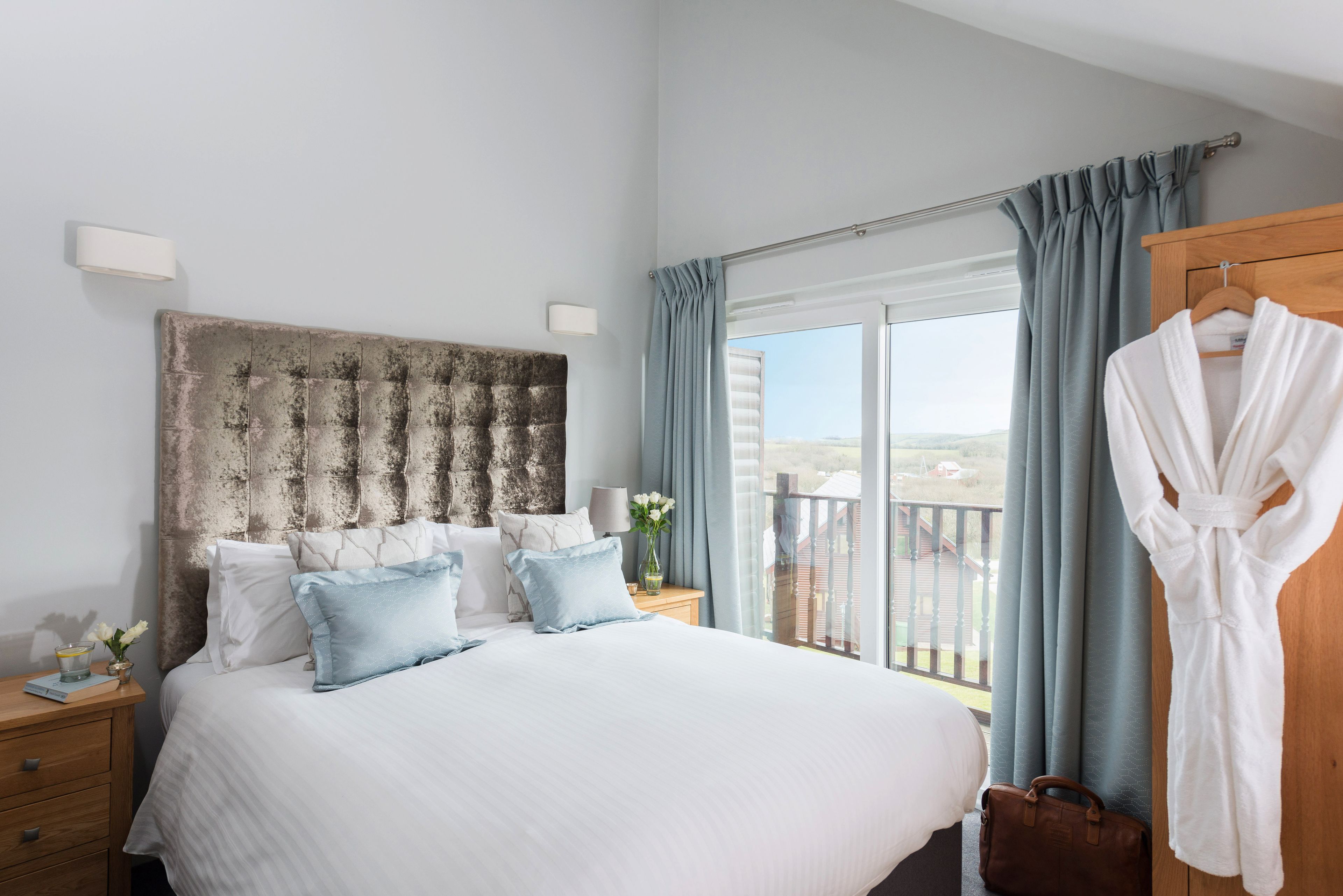 A bedroom with a large bed, a wooden headboard, a nightstand with a lamp, a window with curtains, and a robe hanging on the wall.