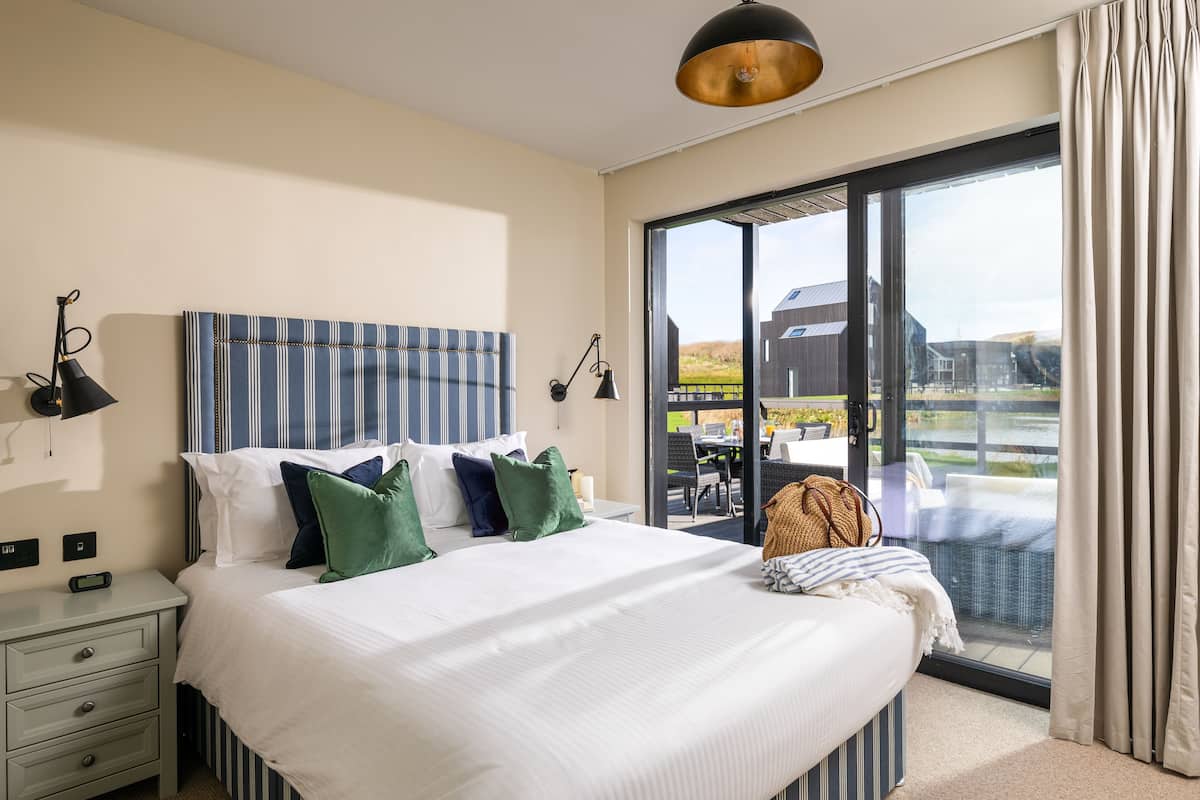 A hotel room with a large bed, a nightstand, a lamp, and a view of the outdoors through a sliding glass door.