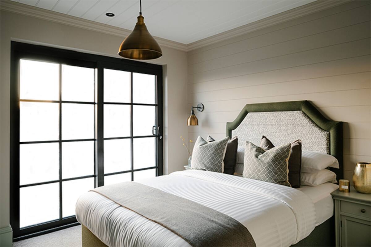 A bedroom with a large bed, a green headboard, a nightstand, a wall-mounted lamp, and a sliding glass door.
