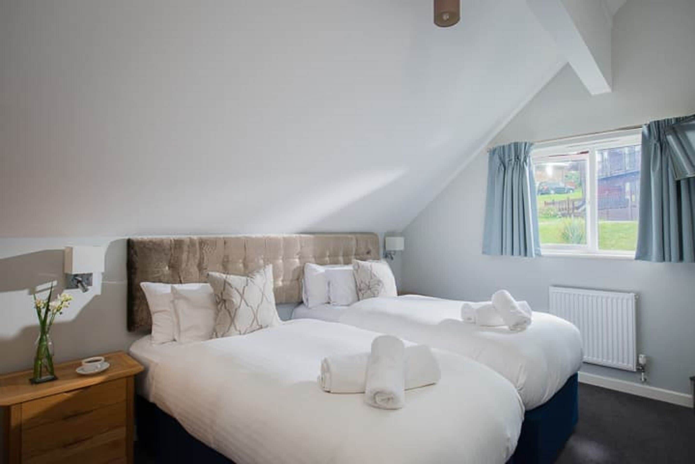 Ein Hotelzimmer mit einem großen Bett, einem Holfnachtkästchen, einer Vase mit Blumen und einem Vorhangfenster.