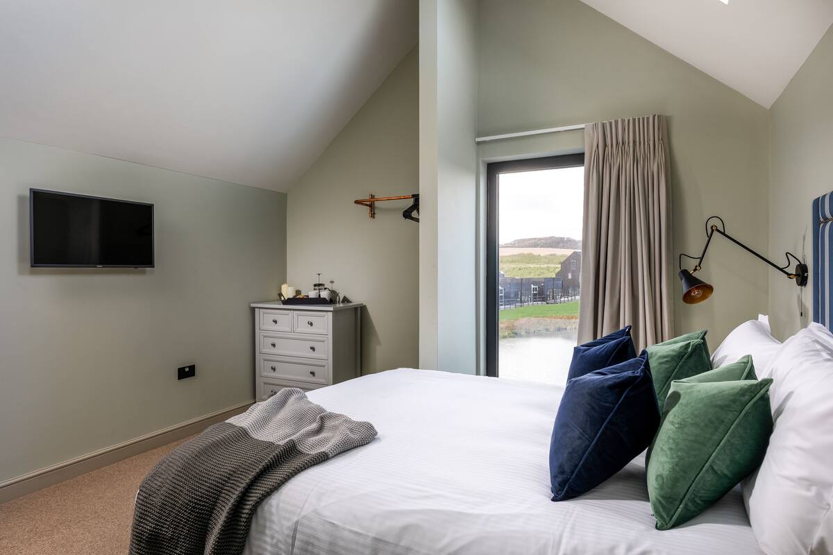 A bedroom with a large bed, a flat-screen TV mounted on the wall, a dresser, and a window with curtains.